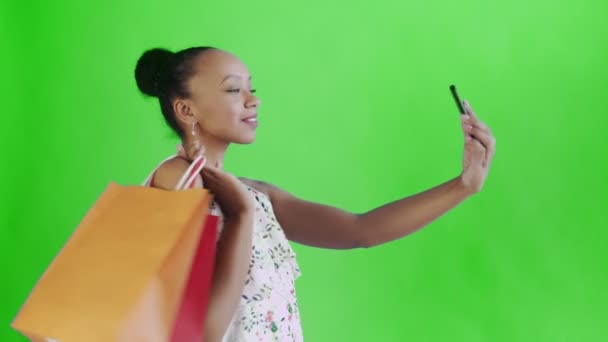 Az afro-amerikai nő bevásárlószatyrokkal szelfit készít a zöld hátteréről a stúdióban. Fehér ruha virágokkal — Stock videók