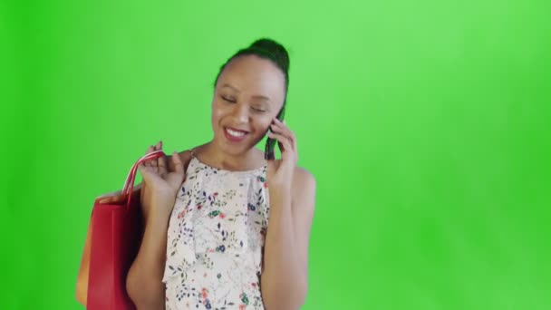 Fiatal gyönyörű afrikai használja a telefont, és tartsa bevásárló táskák zöld háttér stúdióban. Fehér ruha virágokkal — Stock videók