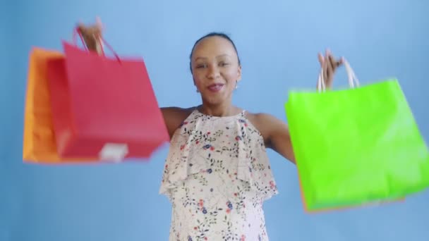 Retrato de mulher afro-americana está dançando com sacos de compras em fundo azul no Studio. Happy Woman Holding Sacos coloridos de compras. Vestido branco com flores — Vídeo de Stock