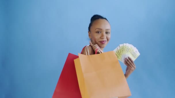 Mulher afro-americana com cartão de dinheiro e sacos de compras. Fundo azul. Vestido branco com flores — Vídeo de Stock