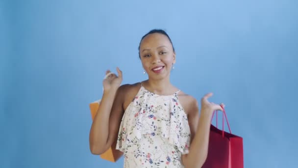 Afrikanische Amerikanerin posiert mit Einkaufstaschen in beiden Händen auf blauem Hintergrund im Studio. glückliche Frau mit bunten Einkaufstaschen. weißes Kleid mit Blumen — Stockvideo