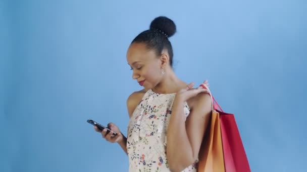 Portrait of african american woman with shopping bags is talking by smartphone on blue Background in Studio. White dress with flowers — Stock Video