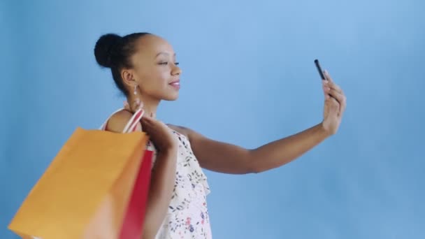 Donna afroamericana con borse della spesa sta facendo un selfie su sfondo blu in Studio. Abito bianco con fiori — Video Stock