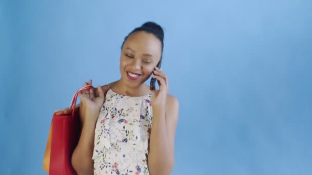 Mladý krásný Afričan pomocí telefonu a držení nákupních tašek na modrém pozadí ve studiu. Bílé šaty s květinami — Stock video