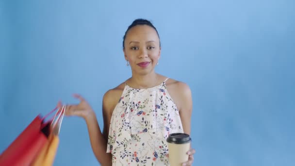 Retrato de una mujer afroamericana con bolsas de compras está bebiendo un café sobre fondo azul en Studio. Vestido JWhite con flores — Vídeos de Stock