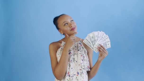 Thoughtful successful african american woman with money on blue background. White dress with flowers — Stockvideo