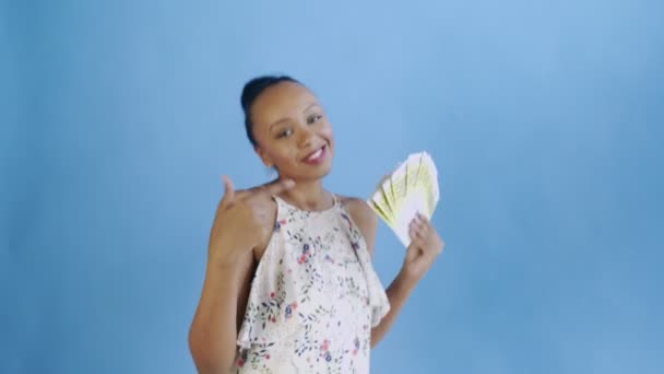 Femme afro-américaine danse avec de l'argent et montrant pouce vers le haut sur fond bleu dans Studio. Robe blanche avec des fleurs — Video