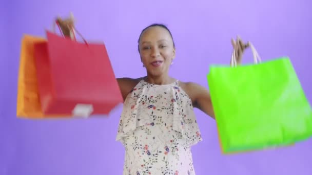 Portrét africké Američanky tančí s nákupními taškami na fialovém pozadí ve studiu. Šťastná žena drží nákupní barevné tašky. Bílé šaty s květinami — Stock video