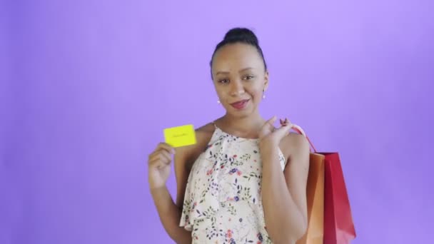 Αφροαμερικανή γυναίκα με πιστωτική κάρτα και πακέτα στα χέρια της. Μωβ υπόκρουση. Λευκό φόρεμα με λουλούδια — Αρχείο Βίντεο