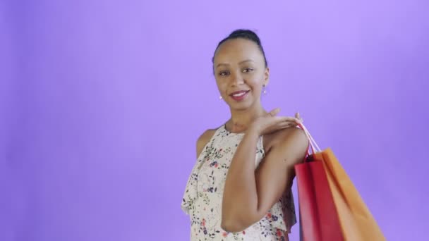 Afroamerykanka pozuje z torbami na fioletowym tle w Studio. Szczęśliwa kobieta trzyma kolorowe torby na zakupy. biała sukienka z kwiatami — Wideo stockowe