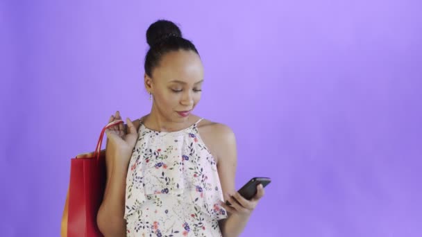 Fiatal gyönyörű afrikai használja a telefont, és tartsa bevásárló táskák lila háttér stúdióban. Fehér ruha virágokkal — Stock videók