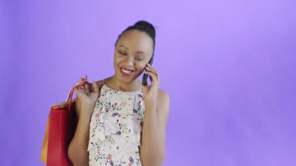 Giovane bella africana utilizzando il telefono e tenendo borse della spesa su sfondo viola in Studio. Abito bianco con fiori — Video Stock