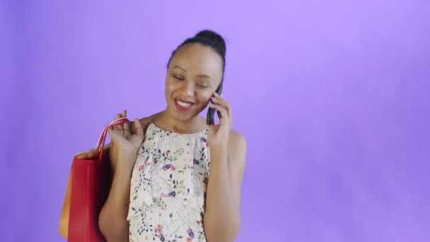 Jovem bela Africano usando telefone e segurando sacos de compras no fundo roxo no estúdio. Vestido branco com flores — Vídeo de Stock