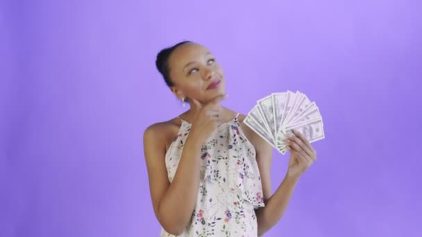 Thoughtful successful african american woman with money on purple background. White dress with flowers — 图库视频影像