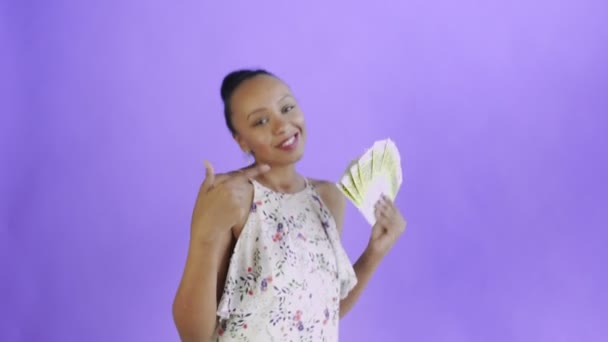 Mujer afroamericana está bailando con dinero y mostrando el pulgar hacia arriba en fondo púrpura en Studio. Vestido blanco con flores — Vídeos de Stock
