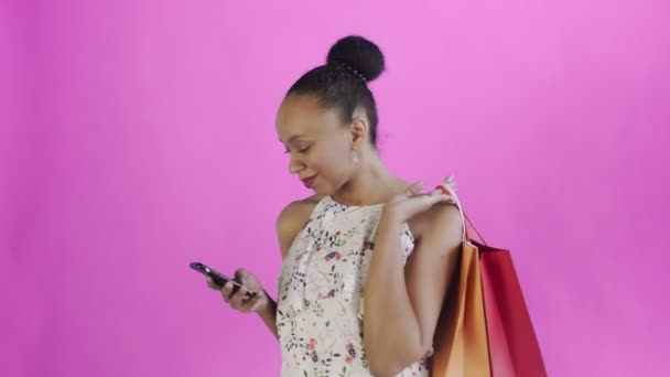 Portrait of african american woman with shopping bags is talking by smartphone on pink Background in Studio. White dress with flowers — ストック動画