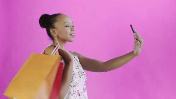 Afrikai amerikai nő bevásárlószatyrokkal szelfit készít rózsaszín hátterről a stúdióban. Fehér ruha virágokkal — Stock videók