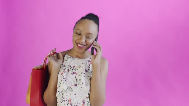 Fiatal gyönyörű afrikai használja a telefont, és tartsa bevásárló táskák rózsaszín háttér stúdióban. Fehér ruha virágokkal — Stock videók