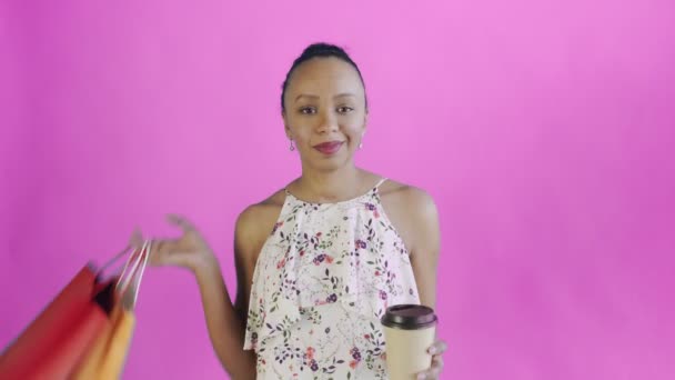 Retrato de una mujer afroamericana con bolsas de compras está bebiendo un café sobre fondo rosa en Studio. Vestido JWhite con flores — Vídeos de Stock