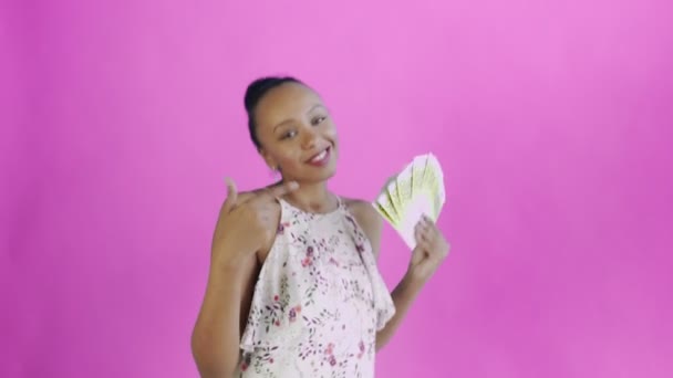 Африканська американка танцює з грошима і показує великий палець на рожевому фоні в студії. Біла сукня з квітами. — стокове відео
