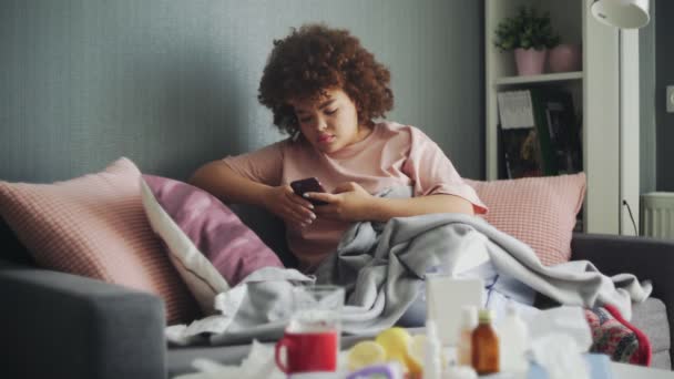 Beteg afro-amerikai lány influenzavírussal a kanapén otthon. Üzenetet ír orvosának, vagy a betegség tüneteit keresi az interneten. — Stock videók