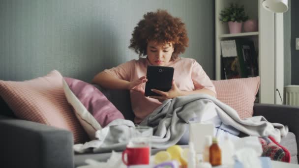 Fille américaine africaine malade avec le virus de la grippe sur le canapé à la maison. Elle utilise une tablette pour rechercher les symptômes de la maladie sur Internet — Video