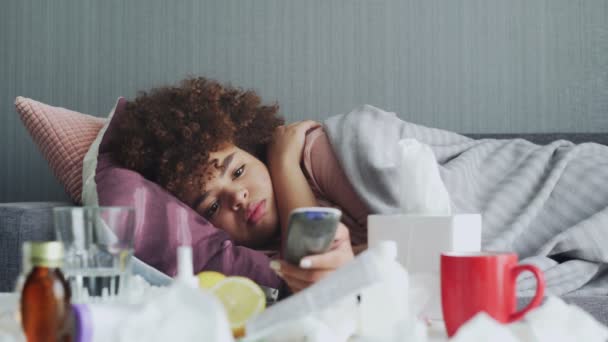 Ziek Afrikaans Amerikaans meisje met griep thuis, ziek jonge zwarte vrouw met koorts kijken tv-show en met behulp van afstandsbediening. — Stockvideo