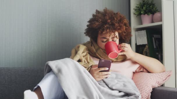 Ziek jong afrikaans amerikaans meisje met behulp van telefoon en het drinken van een hete thee thuis — Stockvideo