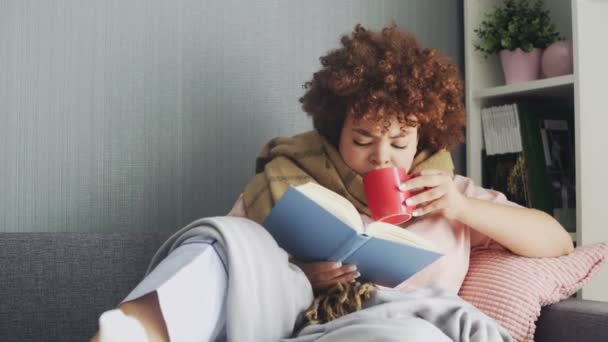 Una mujer tiene un resfriado en casa. Acostado en el sofá con un libro y una taza de té caliente, cubierto con una manta — Vídeo de stock