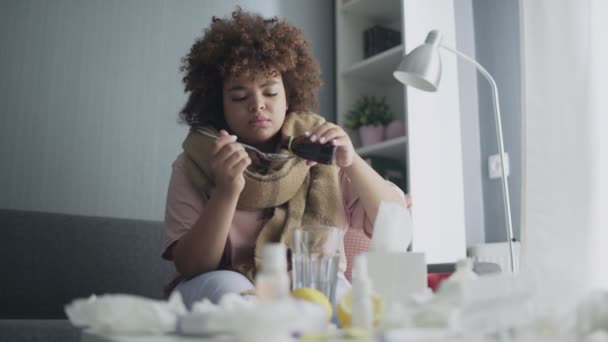 Ziek Afrikaans Amerikaans meisje gieten medicatie of antipyretische siroop om lepel en drinken met water, zittend op de bank. — Stockvideo