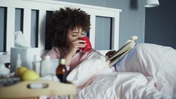 La chica afroamericana enferma tiene un resfriado en casa. Acostado en la cama con un libro y una taza de té caliente — Vídeos de Stock