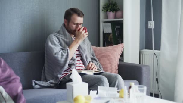 Dospělý nemocný muž sedí na pohovce pokrytý šedými kostkami, čte knihu a pije horký čaj — Stock video