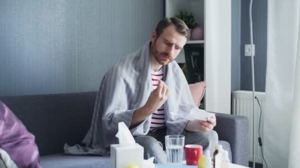Взрослый больной мужчина сидит на диване и принимает таблетки с водой — стоковое видео