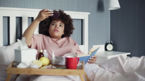 Enfermo joven afroamericano chica está acostado con un libro en la cama y el uso de teléfono para hacer un video o historias — Vídeos de Stock