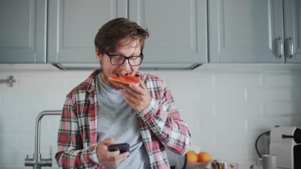 Young man eating toast with strawberry jam and using phone — Stock Video