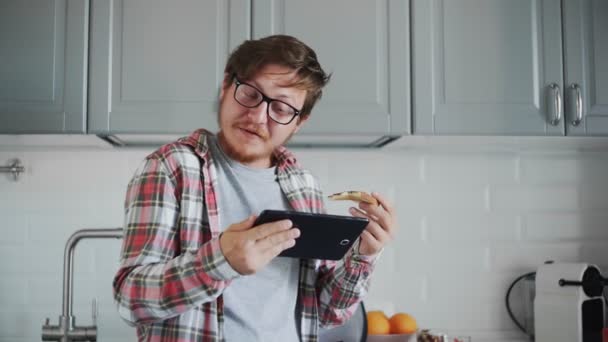 Jovem com tablet comendo torrada com pasta de chocolate. Homem assistir a um vídeo engraçado no tablet e rir — Vídeo de Stock