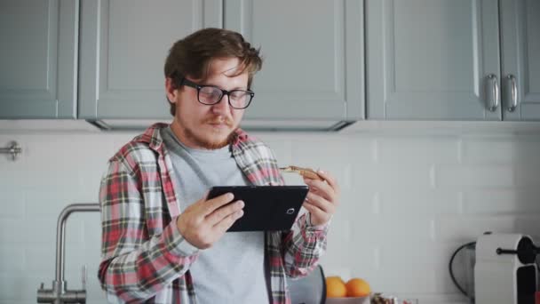 タブレットの若い男はチョコレートペーストでトーストを食べる。男見ますA面白いビデオオンタブレットと笑い — ストック動画