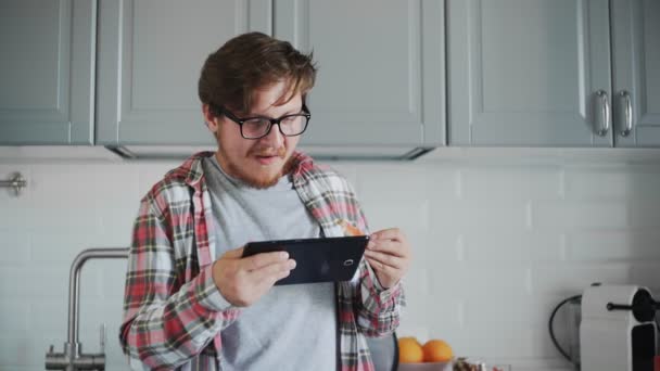 Mladý muž s tabletou jí tousty s jahodovou marmeládou. Muž sledovat legrační video na tabletu a smát — Stock video