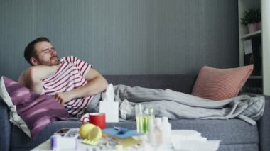 Evdeki kanepede grip olmuş hasta bir adam yatıyor. Soğuk termometreyle ateşini ölçen hasta bir genç.