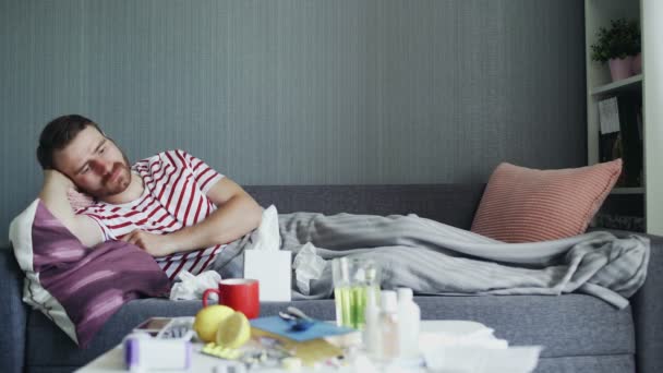 Άρρωστος άντρας με γρίπη ξαπλωμένος στον καναπέ στο σπίτι. lll νεαρός άνδρας με κρύο θερμοκρασία ελέγχου με θερμόμετρο — Αρχείο Βίντεο