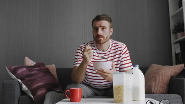 Hombre joven viendo la televisión durante el desayuno sentado en el sofá en la habitación — Vídeos de Stock