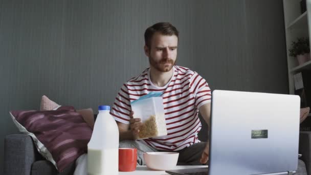 Skäggig man förbereder frukost och använder en bärbar dator — Stockvideo