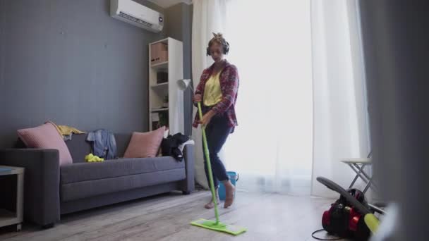 Kulaklıklı kadın evde paspasla yerleri yıkıyor ve dans ediyor. — Stok video