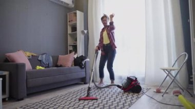 Evde elektrikli süpürgeyle halıyı temizlerken oturma odasında dans eden neşeli genç bir kadın..