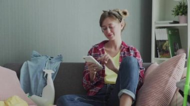 Güzel genç bayan temizlik yaptıktan sonra kanepede dinleniyor ve telefonu kullanıyor.
