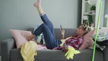 Genç bayan evi temizledikten sonra telefon edip kanepede dinleniyor.