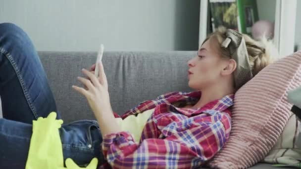 Young woman is using phone and resting on the sofa after cleaning house — Stock Video