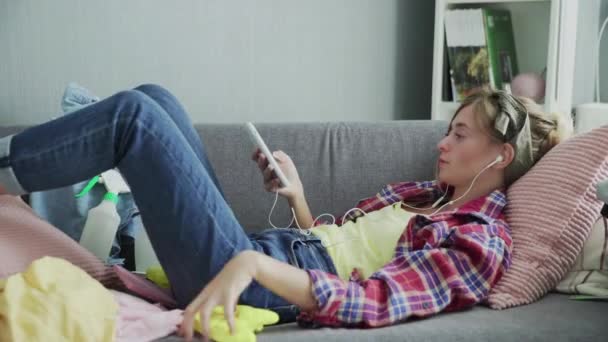 Молодая женщина слушает музыку и отдыхает на диване после уборки дома — стоковое видео