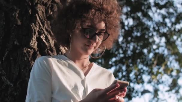 Närbild av lockig kvinna använder mobiltelefon i den offentliga parken. — Stockvideo