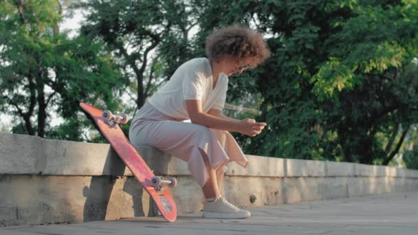 Портрет хіпстерської молодої жінки зі скейтбордом і телефоном, що сидить у парку — стокове відео
